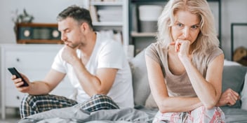 angry looking woman sitting at end of bed while a man looks at his phone behind her